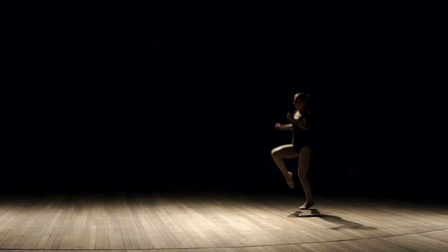 A flexible girl in a black swimsuit makes a gymnastic somersault on stage on a black background, slow motion. Girl doing gymnastic flip. Acrobatic flip. Ballet. Dancer.