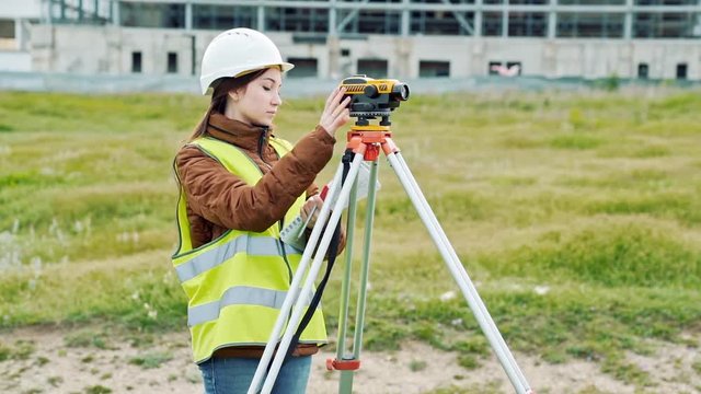 A young woman surveyor in work clothes and helmet adjusts the equipment and produces calculations on the construction site. The concept of landscape design, geodesy