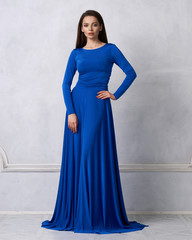 Young woman with long brunette hair dressed in elegant evening blue maxi dress with long sleeves posing in studio. Gorgeous female model standing against white wall decorated with classic moldings. - obrazy, fototapety, plakaty