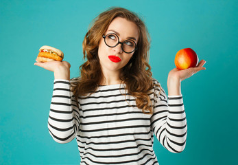 Portrait of young beautiful blond woman wearing glasses  holding red apple in one hand and burger in another over blue background