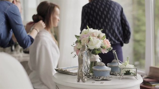 Brides morning shallow dof. Light closeup of table with bridal bouquet and rings in focus and two young professional stylists working on girl image fixing hair checking make-up at defocused background