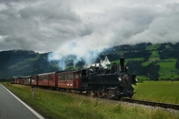 Fototapeta na wymiar Retro locomotive with smoking engine moving forward in Alps mountains. Scenic view on the vintage train against the backdrop of the forest and beautiful hills in Europe