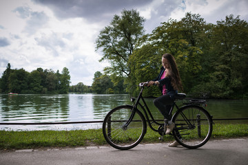 Young woman riding a bicycle in the park on the background of trees and lake. Trendy active girl spends time on the nature. Side view.