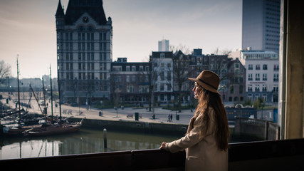 Young woman is standing on the balcony and looking on old marina and city center in Rotterdam, Netherlands. Elegant female tourist explore city in sunny day. Trendy girl enjoys traveling in Europe