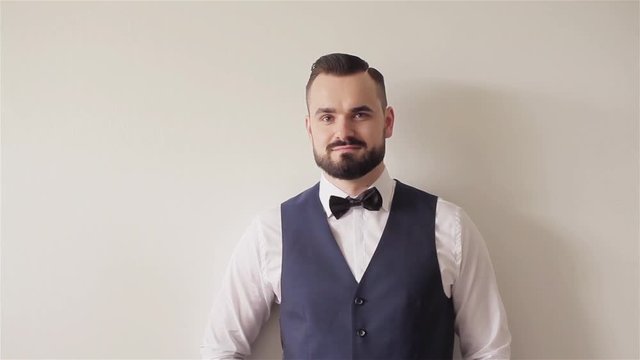 Portrait of 30s bearded man with bowtie stands posing at white wall looks at camera smiling. Barber shop client with hair gelled. Appearance apparel attire accessories and classic male fashion details