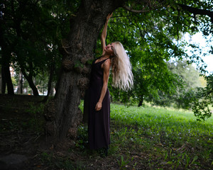 Young slim woman stands under big tree and relaxes in the park. Athletic blonde girl with beautiful...