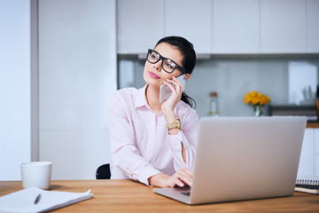 Young woman answering phone call while working on laptop in home office