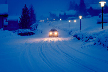 Car driving in fresh snow on an early morning in the ski town called Zinal. The street and car lights are lightning the snow covered road.