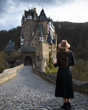 Young elegant woman with bottle of wine stands on the road leading to fabulous castle. Incredible medieval place. Sunny rays falling on the hair of female traveler. Trendy girl is traveling in Europe