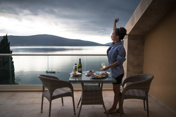 Young happy woman is standing on the balcony of apartment near the table with food and drinks and waving to somebody. Smiling girl with glass of wine is looking on the beautiful sea and mountains
