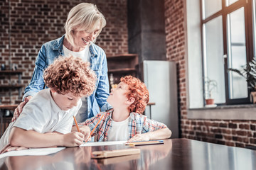 Studying at home. Delighted nice red haired boy sitting at the table and looking at his grandmother while studying at home together with her