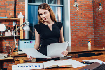 Serious woman reading papers studying resumes standing at work desk in stylish office