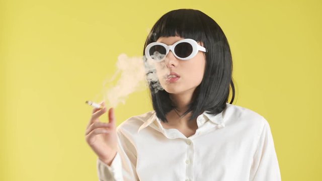 Portrait of sassy girl 20s in white vintage outfit and sunglasses smoking cigarette with disgust, over yellow background in studio. Concept of emotions