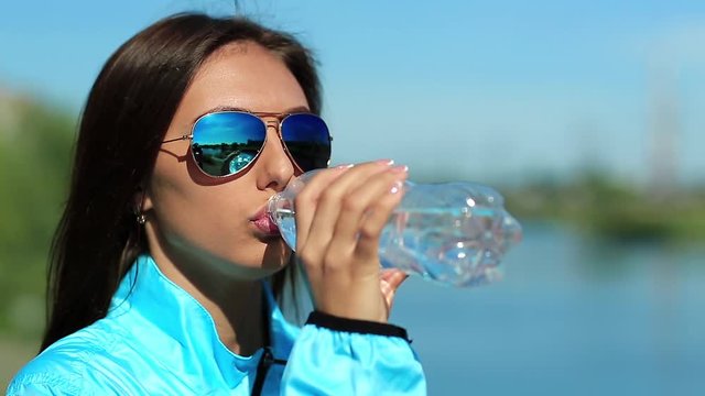 Sportswoman drinks water. Attractive girl in sunglasses drinks clean mineral water from bottle. Beautiful woman in blue sports jacket drinks water and looks around