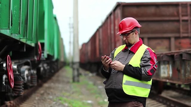 Railwayman uses smartphone near goods train. Railway employee in working clothes on railway line holds in hands mobile phone, works with smartphone on freight station