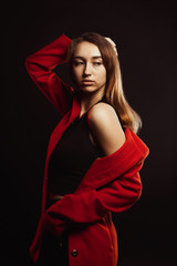 Stylish brunette woman with natural makeup and shiny hair posing in trendy red coat at studio