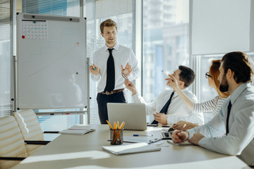 Aim high. Pleasant young boss standing near the whiteboard with a graph on it and explaining their new project objectives to his colleagues while they asking questions