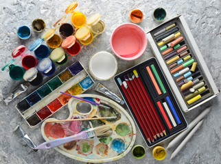 Obraz premium Artistic tools for drawing paintings on a gray concrete background. Palette, gouache, oil paint, brushes, colored crayons, pastel, colored pencils. Top view..