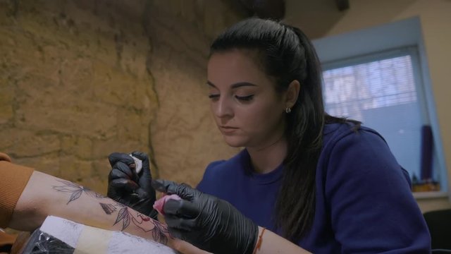 brunette female tattooer in violet clothing making tattoo on the man's arm of her client with gun in studio. Young business woman in workshop. 4k