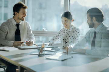 Friendly ambience. Upbeat young colleagues sitting at the table next to their boss and joking together with him while having a meeting