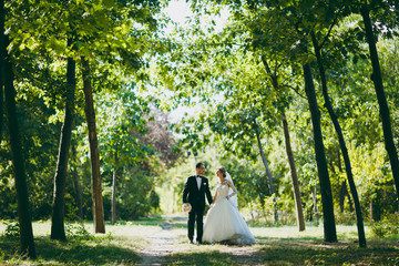 Fototapeta premium Beautiful wedding photosession. The groom in a black suit with bouquet and his bride in a white lace dress with a long plume and veil smile and walk along the path in large green garden on sunny day