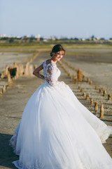 Fototapeta na wymiar Beautiful wedding photosession. Young cute elegant shapely bride in white lace dress with a long plume waving in the wind on a walk along the coastline against a beautiful nature landscape