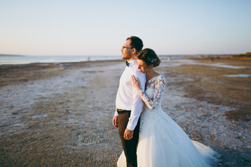 Fototapeta na wymiar Beautiful wedding photosession. Handsome unshaved groom in white shirt and young cute bride in white lace pattern dress with exquisite hairstyle looking into distance on walk along coastline sunset