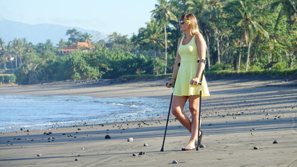 Lady in a yellow sundress with crutches looking into the distance on sandy beach
