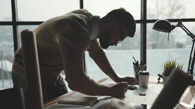 Engineer works in a bright office with a large window, concentrates and draws blueprints. Workplace of an architect or designer: loft style, minimalistic interior, drawings and coffee on the table.