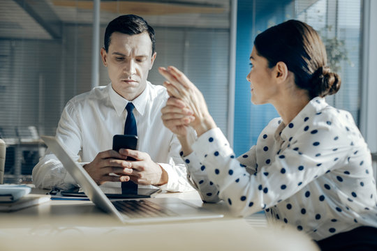 Distracting from topic. Handsome young man sitting at the table next to his female colleague and checking his phone, distracting himself from the work-related discussion with her
