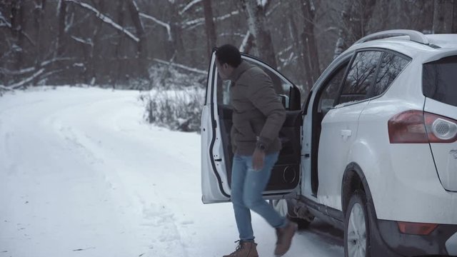 Black man walking out of stopped car confused with breakdown looking around in winter woods.