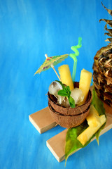 Ingredients for pina colada cocktail. Ice cubes and pineapple pieces served in a coconut half decorated with an umbrella and straw on blue background
