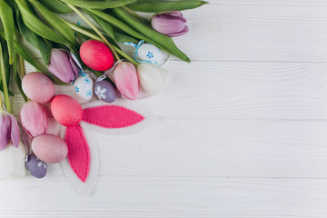Easter composition with colored eggs, rabbit ears and tulips on a white wooden background.