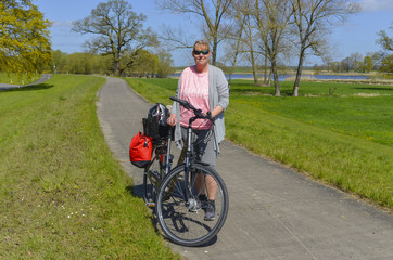 Obraz premium Woman standing on a bikeway beside her bike in rural surrounding near the Elbe River in the Lueneburg Heath at a sunny day with blue sky - landscape photo