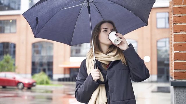 Thoughtful attractive young woman wearing a gray coat is standing under an umbrella and drinking coffee. Handheld slow motion medium shot