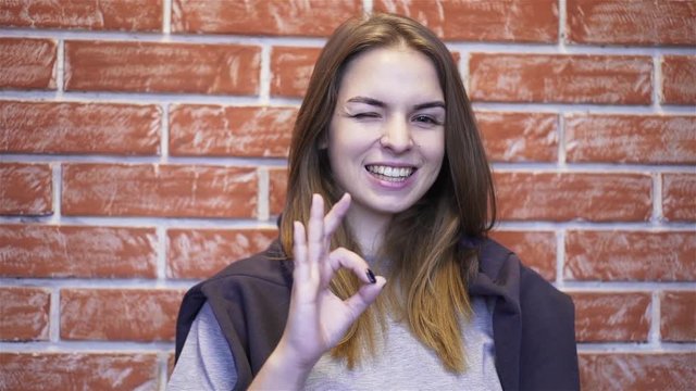 Beautiful young woman showing an ok sign standing in a building corridor with brick walls. Handheld slow motion close up shot