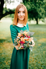 Young happy red haired girl with bouquet of summer flowers enjoying inner peace in green dress at nature. Little child playing in wonderland park. Pretty woman portrait outdoor. Lovely smiling face.