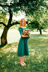 Young happy red haired girl with bouquet of summer flowers enjoying inner peace in green dress at nature. Little child playing in wonderland park. Pretty woman portrait outdoor. Lovely smiling face.