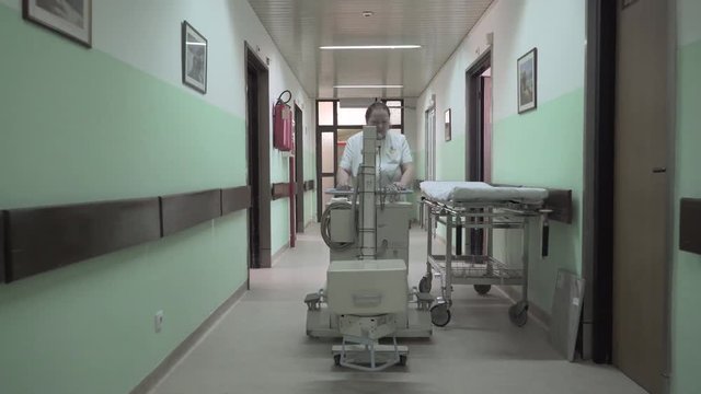 Mature female medic technician pushing medical device and passing through the hallway of hospital ward, nurse moves clinic machine on department, concept medicine, steadicam, tracking, gimbal shot.