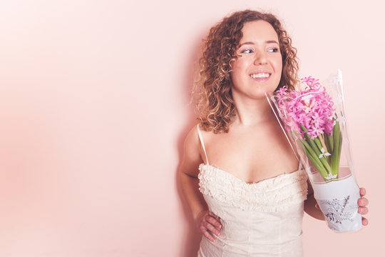 Beautiful curly woman in white dress with hyacinth flowers in hands on a light pink background. Hello spring concept.
