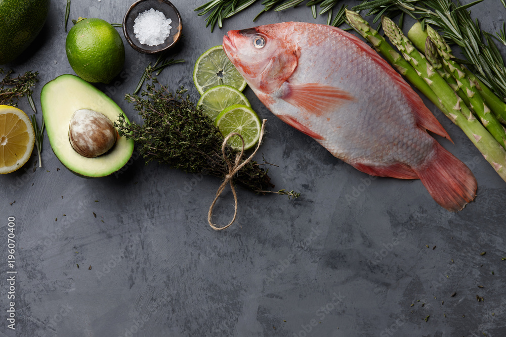 Poster Raw red tilapia fish cooking with herbs, spices, green asparagus, avocado, lemon and lime on gray stone background, top view