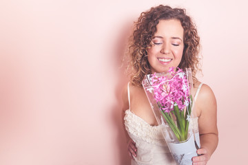 Beautiful curly woman in white dress with hyacinth flowers in hands on a light pink background. Hello spring concept.