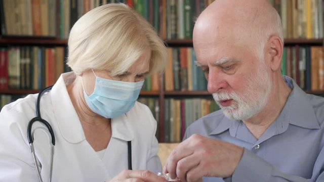 Female professional doctor in white coat and medical mask at work. Senior woman physician treating senior male patient at home giving flu pill. Patient taking medicine with water in glass