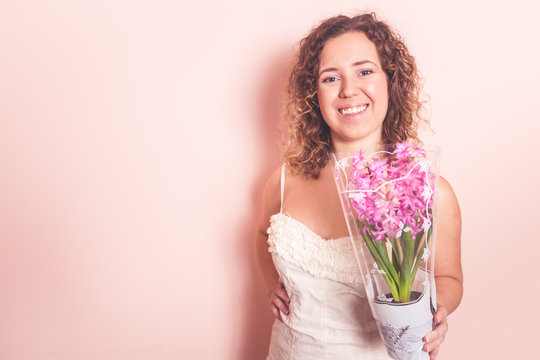 Beautiful curly woman in white dress with hyacinth flowers in hands on a light pink background. Hello spring concept.