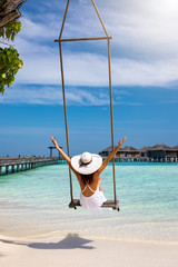 Elegante Urlauberin in weißem Kleid genießt ihren Urlaub auf einer Schaukel am tropischen Strand