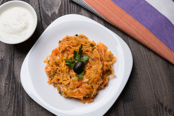 stewed cabbage in a white plate on a wooden table, sour cream and a towel