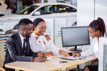 happy African couple and female seller sit at table and make a deal for sale of car