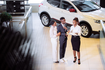 High angle view of african car saleswoman standing at dealership telling about features of car to african couple customers