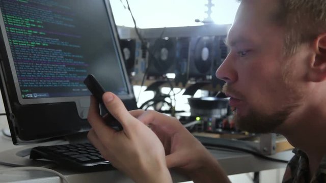 Young man working on mining farm and watching bitcoin currency growth with smartphone.