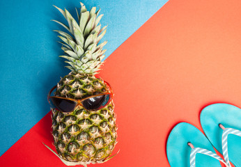 top view of hipster pineapple with sunglasses and blue flip-flops on colorful blue and red...
